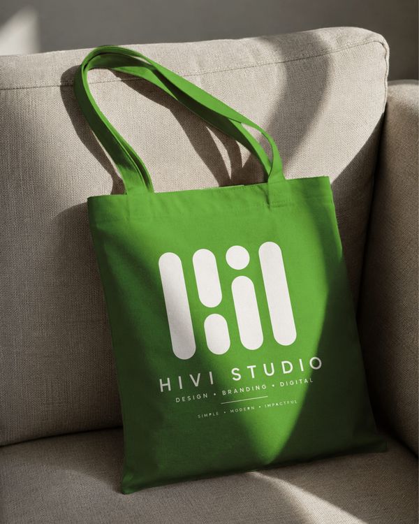 Green canvas tote bag mockup displayed on a sofa with natural lighting.