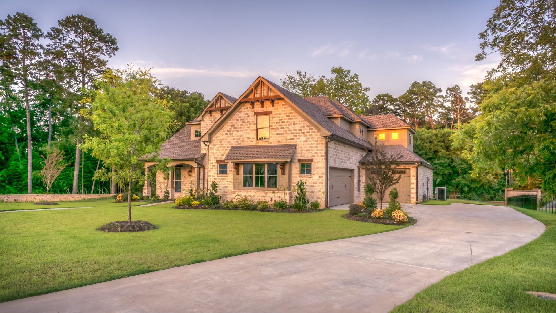 Two-story stone house with a curved concrete driveway, lawn, and pine trees at dusk, with no people, logos, or text