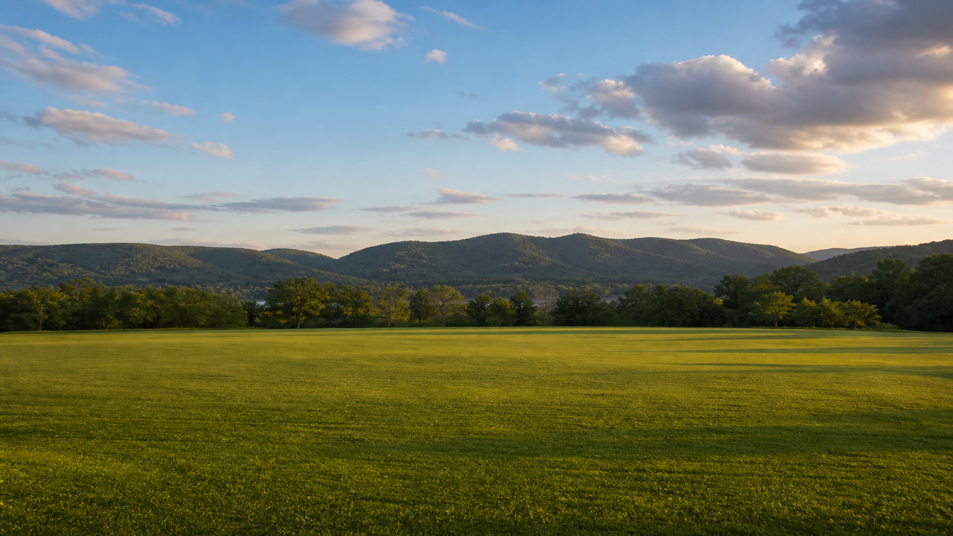 Empty green park and tree line under a late-afternoon sky, with ridgelines beyond and no people, logos, or text