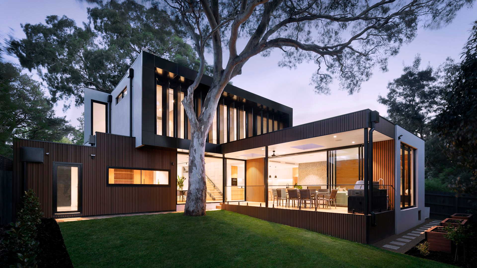 Modern wood-and-glass residence at dusk with interior lights on, empty backyard, no people or text