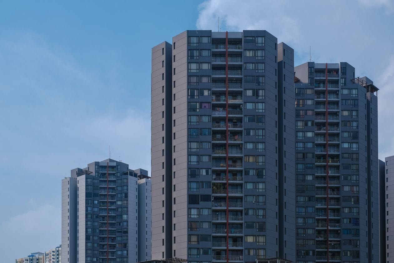 Photoreal low-angle view of a modern mid-rise apartment building with dark cladding and balconies against a blue sky, no people, logos, or text
