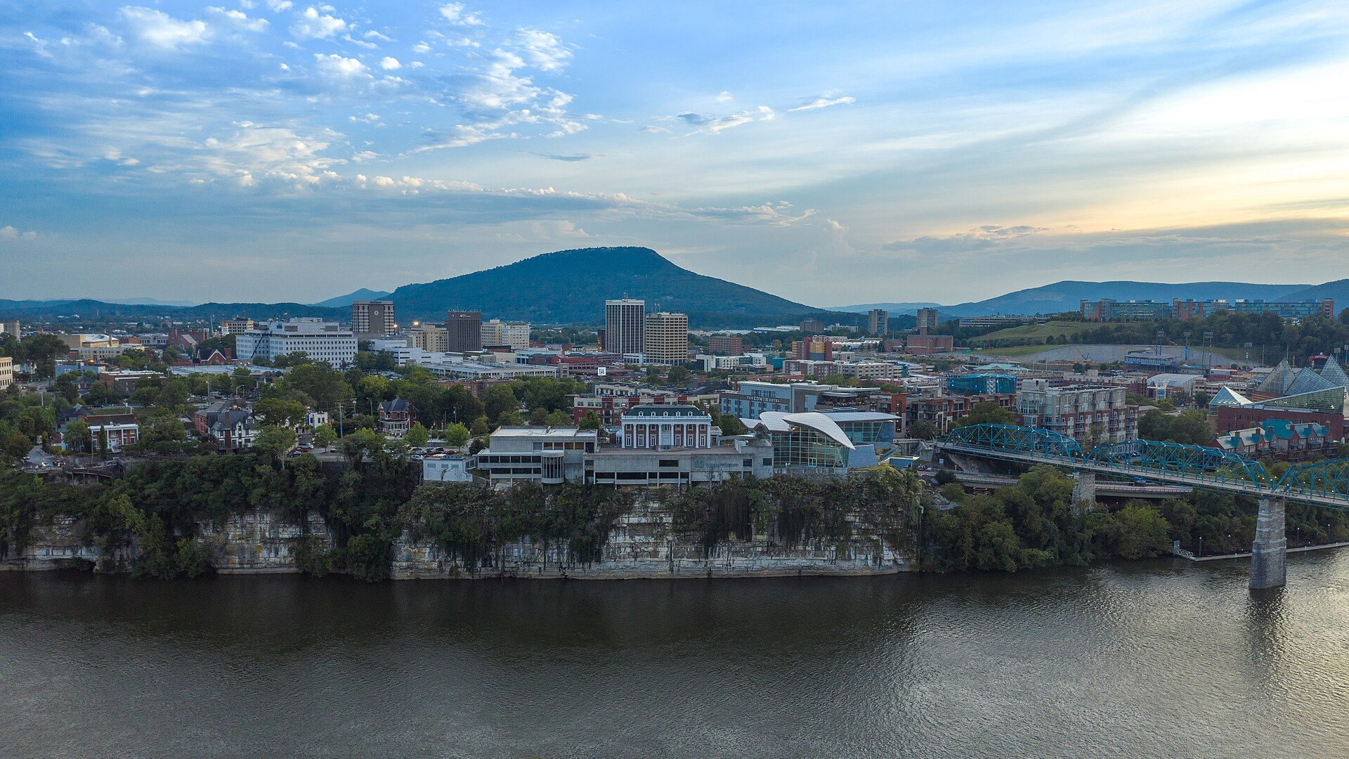 Photoreal view of a riverfront city skyline with a bridge, hills and low clouds over the water, no people, logos, or text