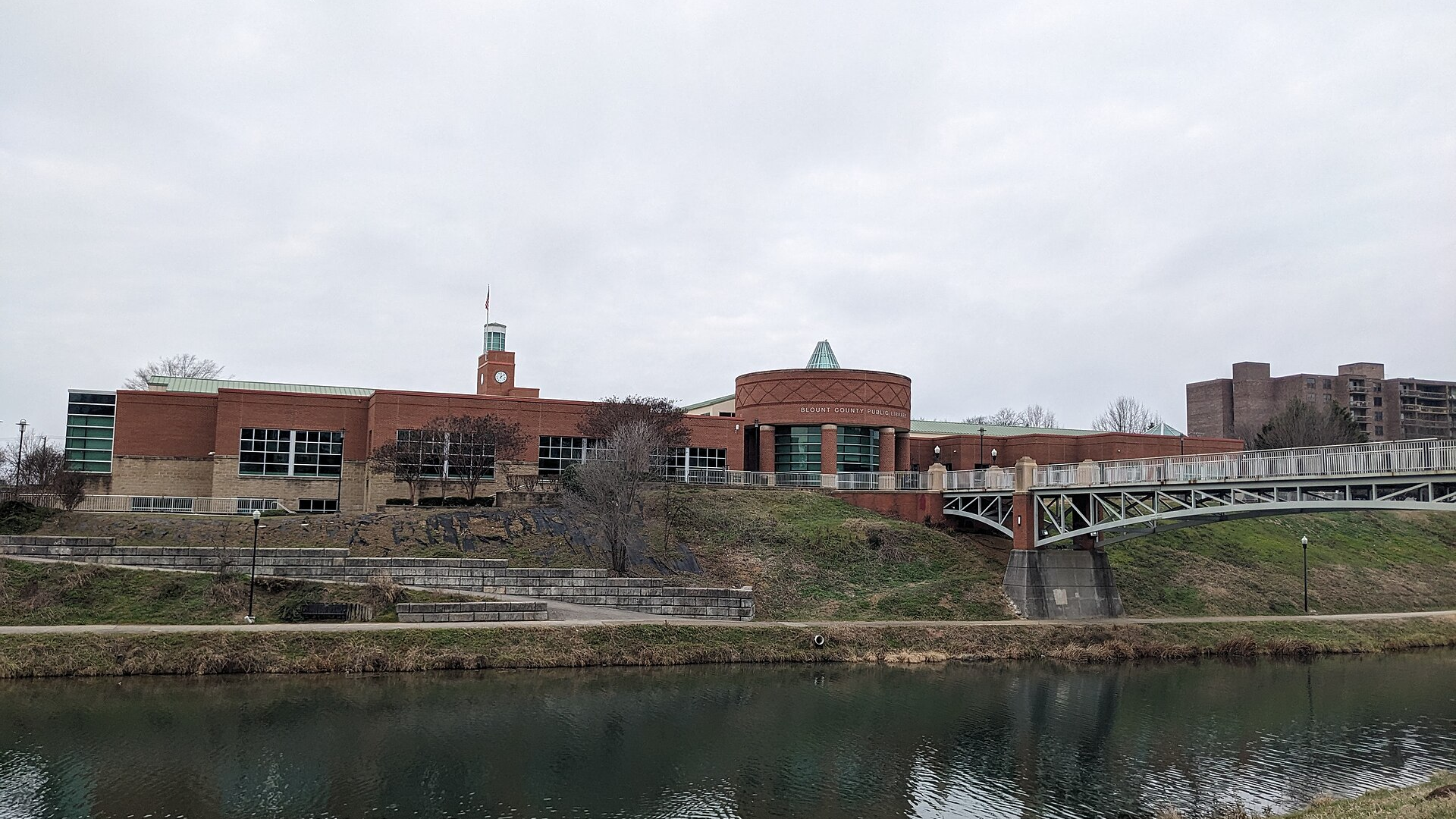 A red-brick public building with a clock tower and a footbridge beside a calm greenway pond under an overcast sky