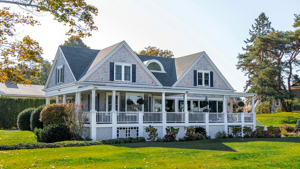 Empty shingled house with a wraparound porch and lawn under a clear sky, with no people, logos, or overlay text