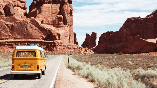 Yellow vintage van on a two-lane desert highway toward red rock cliffs, no identifiable people