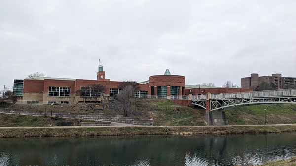 A red-brick public building with a clock tower and a footbridge beside a calm greenway pond under an overcast sky