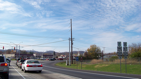 Traffic on a four-lane road in Elizabethton, Tennessee, at a junction marked with US 19E, US 321 and State Route 67 signs, with shops and hills behind