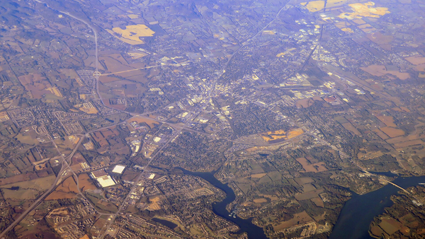 An aerial view of a spread-out town with neighborhoods, roads, and farm fields beside a winding lake