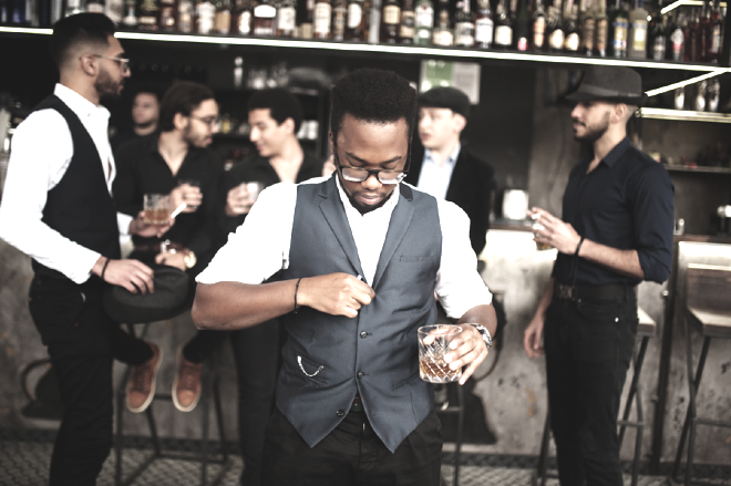 A man in glasses buttons his vest, with a drink in his hand, in a pub.