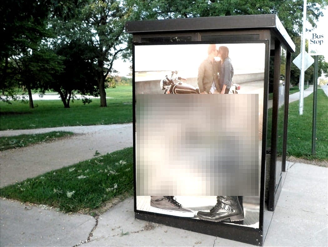 A bus shelter ad display sits beside a quiet campus path and green lawn, with the ad content redacted.