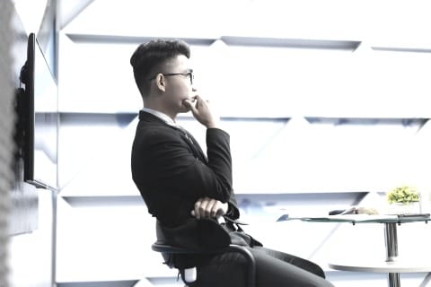 A person in a suit sits sideways on a stool in a bright modern office, hand to chin in a thoughtful pose beside a small table with a plant.