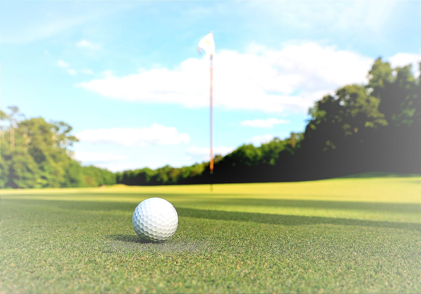 a golf ball sitting on top of a green golf course green with a flag to mark the hole nearby, trees in the background