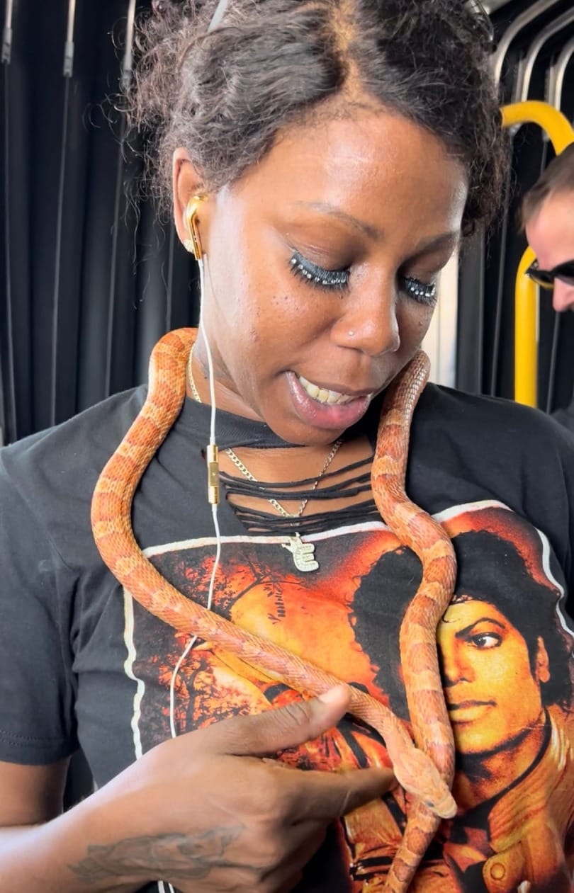 Woman on a New York City metro wearing a corn snake around her neck and shoulders, looking down as the snake stretches across a Michael Jackson T-shirt.