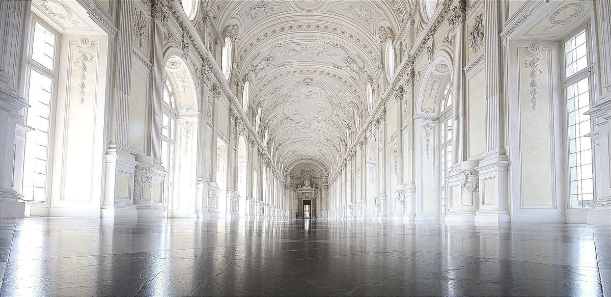 A long, ornate, sunlit hall with high arched ceilings, tall windows, and a polished floor leading toward a distant doorway.