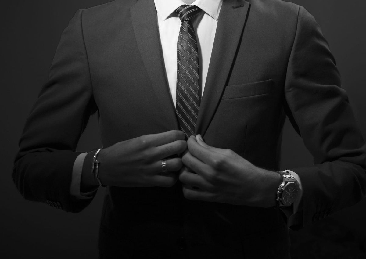 Black-and-white close crop of a man in a dark business suit adjusting his tie, with his face out of frame.