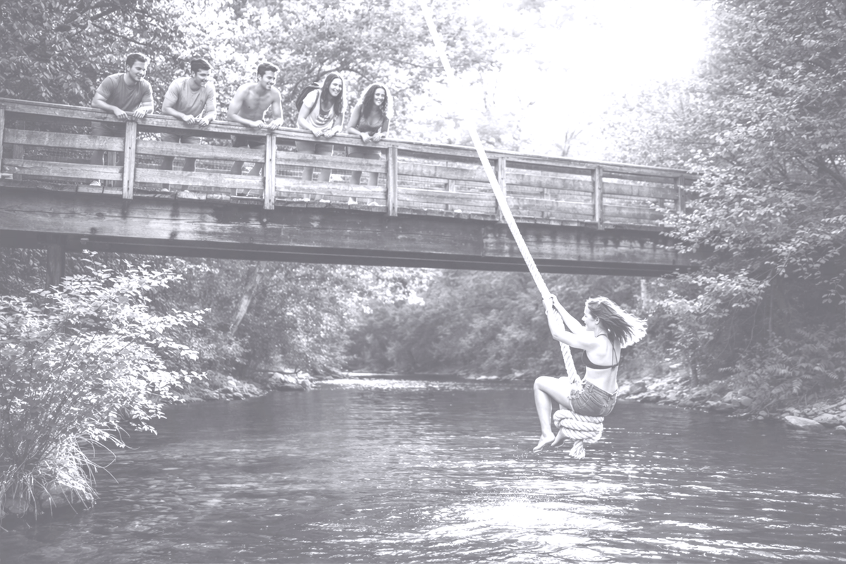 A teenage girl sitting on a rope swing at the high point of its arc above a river, watched by friends standing on a wooden bridge on a summer day.