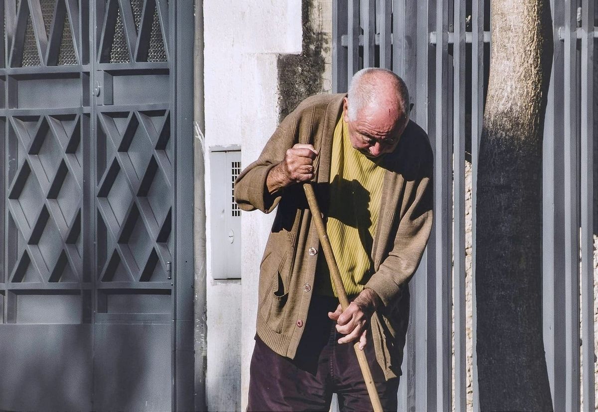 An older man sweeping near a gate and wall, head bent over his broom.