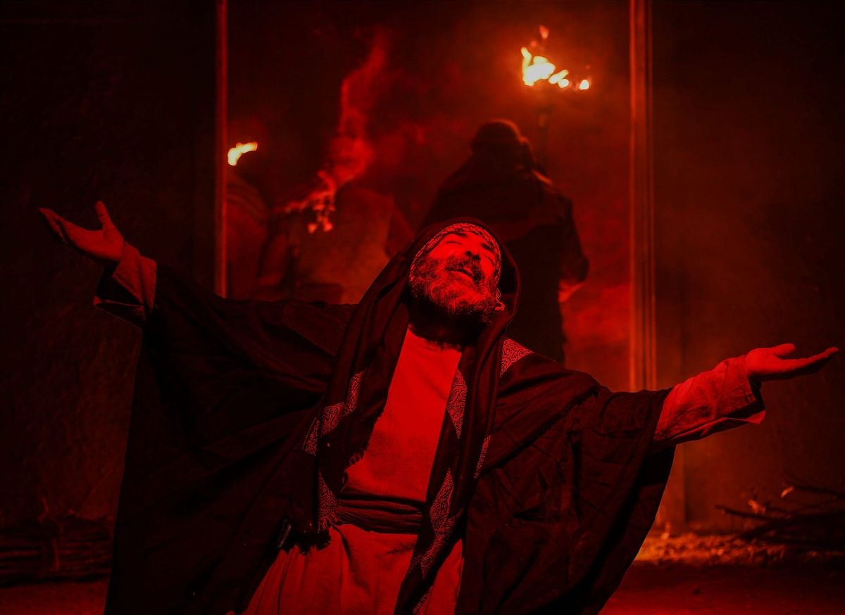 A red-lit, robed figure stands with arms open before fire and shadow, suggesting the theatrical allure and false authority of a cult leader. Photo by Hossein Nasr / Unsplash