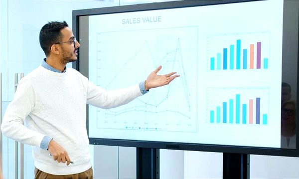 A person gestures toward a large screen displaying charts and bar graphs labeled “Sales Value.”