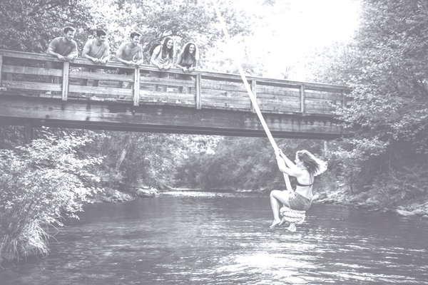 A teenage girl sitting on a rope swing at the high point of its arc above a river, watched by friends standing on a wooden bridge on a summer day.