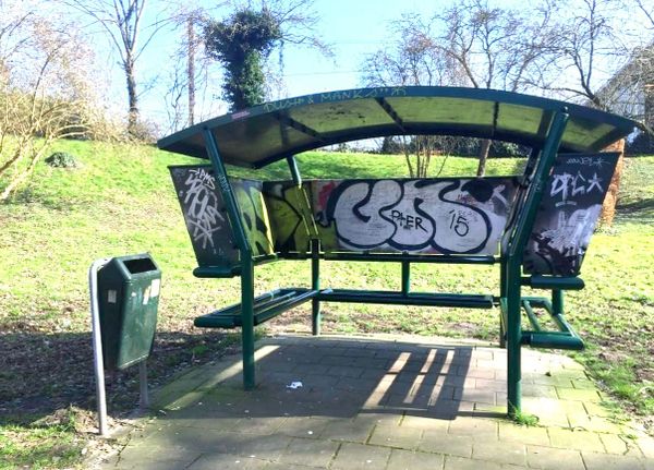 A small covered outdoor shelter with benches, graffiti, and a trash can nearby, set in a grassy neighborhood area. The structure serves as a public hangout space for teenagers.