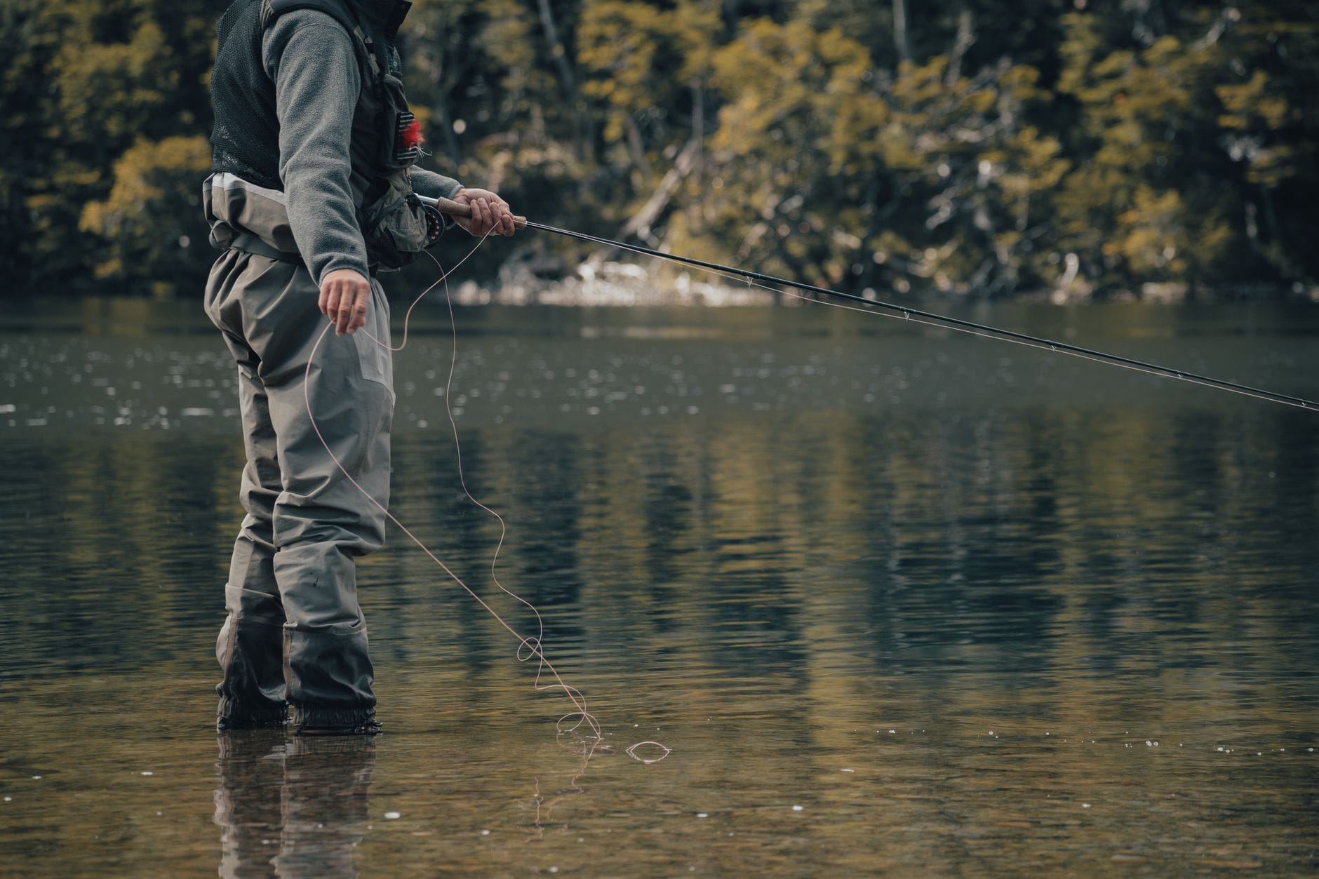 A man stands in reflective water while fly fishing in a serene lake surrounded by trees.