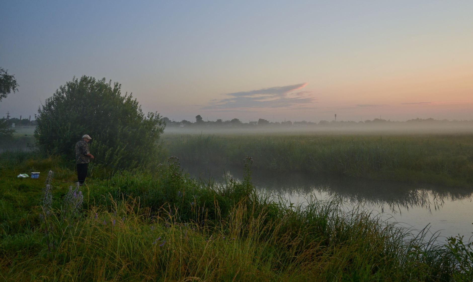 A lone fisherman casting at dawn in a foggy, lush riverside landscape.