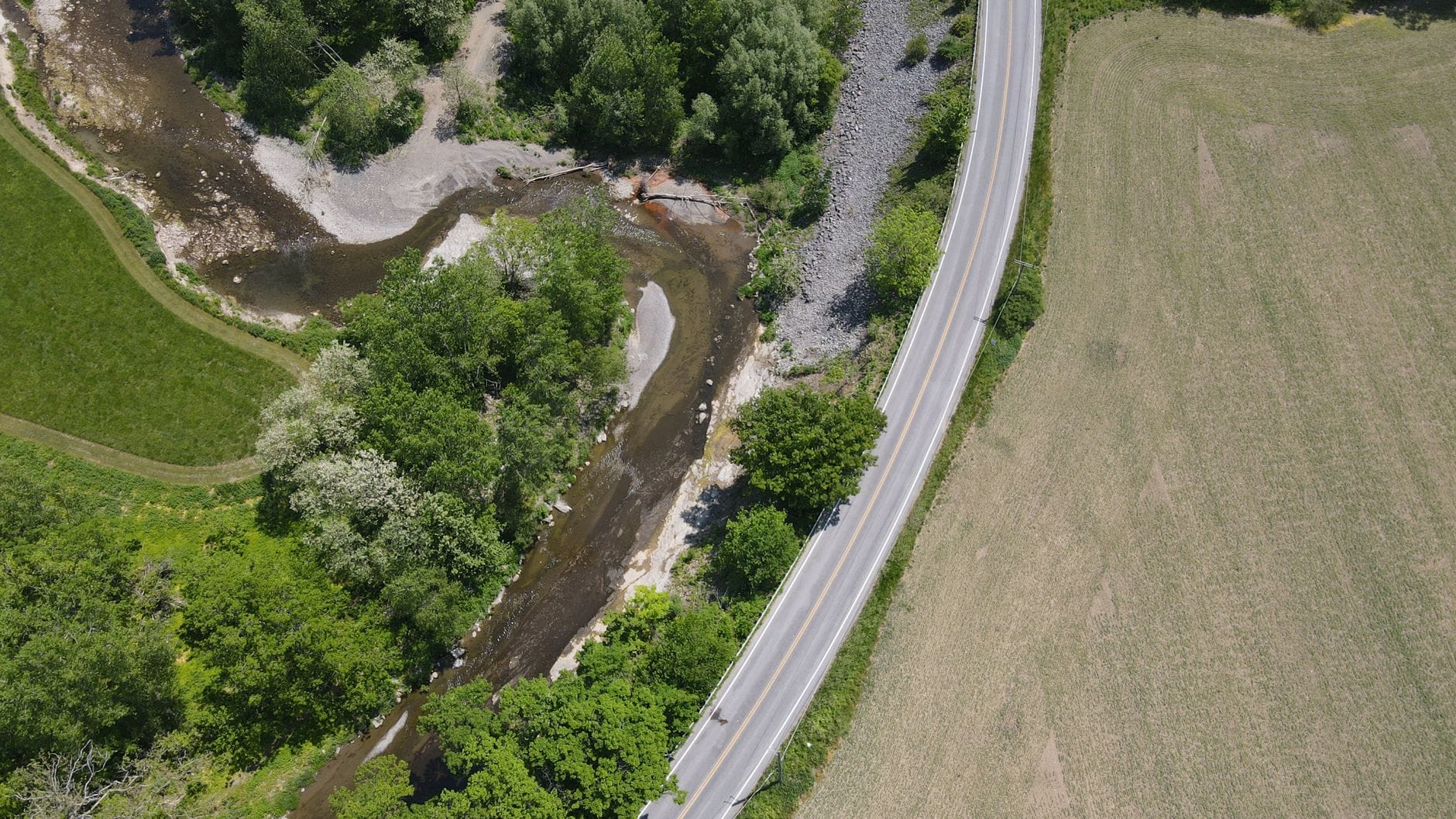 Aerial imagery showcasing a winding rural road beside a flowing river amidst lush green fields during summer.
