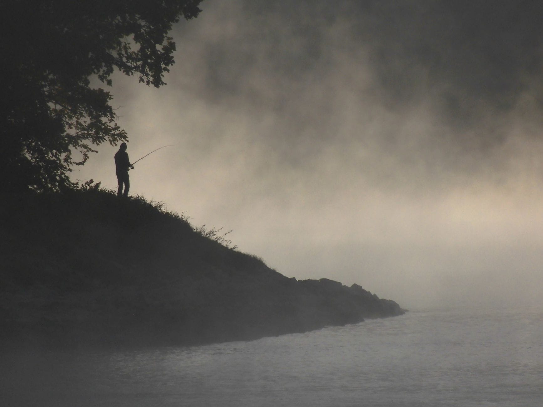 A lone angler silhouetted against misty riverbank at dawn, capturing a serene fishing moment.