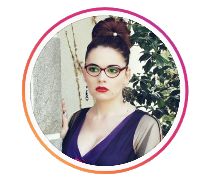 Laura Badler, wearing a blue dress with sheer sleeves and red glasses, looks off to the left in a portrait