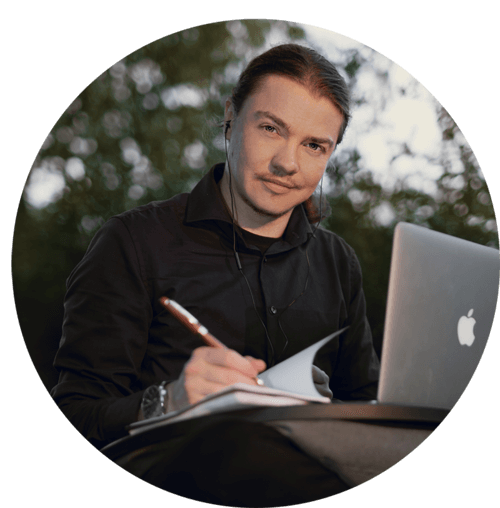 A portrait of Angel Pretot, sitting in nature with his laptop, looking at the camera and using  a pen and notebook with his right hand.