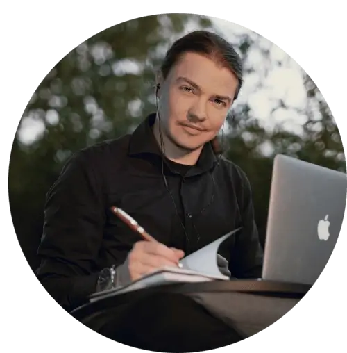A portrait of Angel Pretot, sitting in nature with his laptop, looking at the camera and using  a pen and notebook with his right hand.