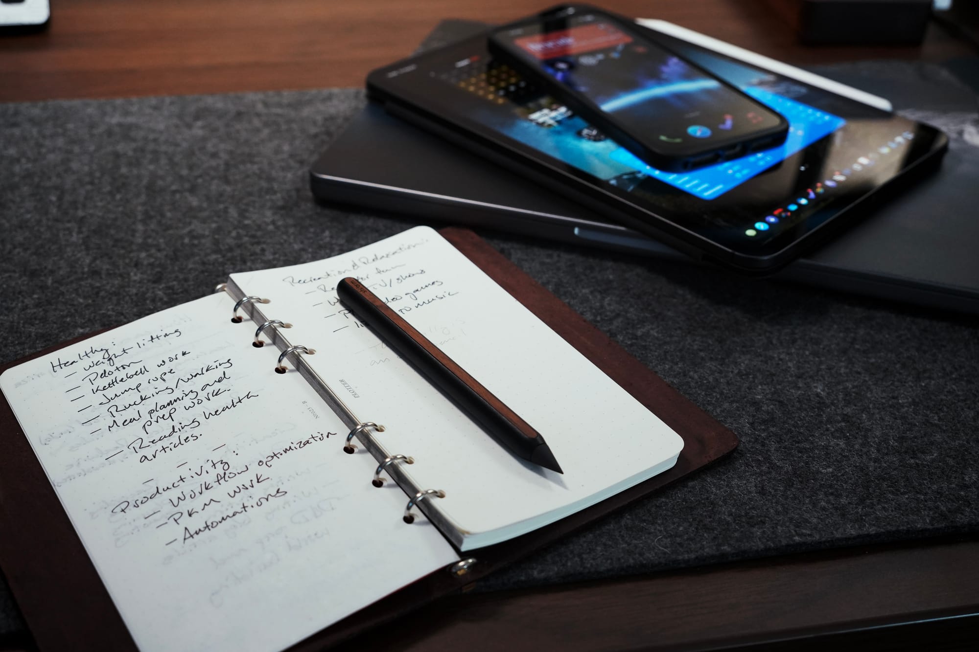 An open notebook with handwritten notes and a black pen resting on its pages, placed on a desk. In the background, a stack of digital devices—a smartphone, tablet, and laptop—contrast with the analog writing tool.