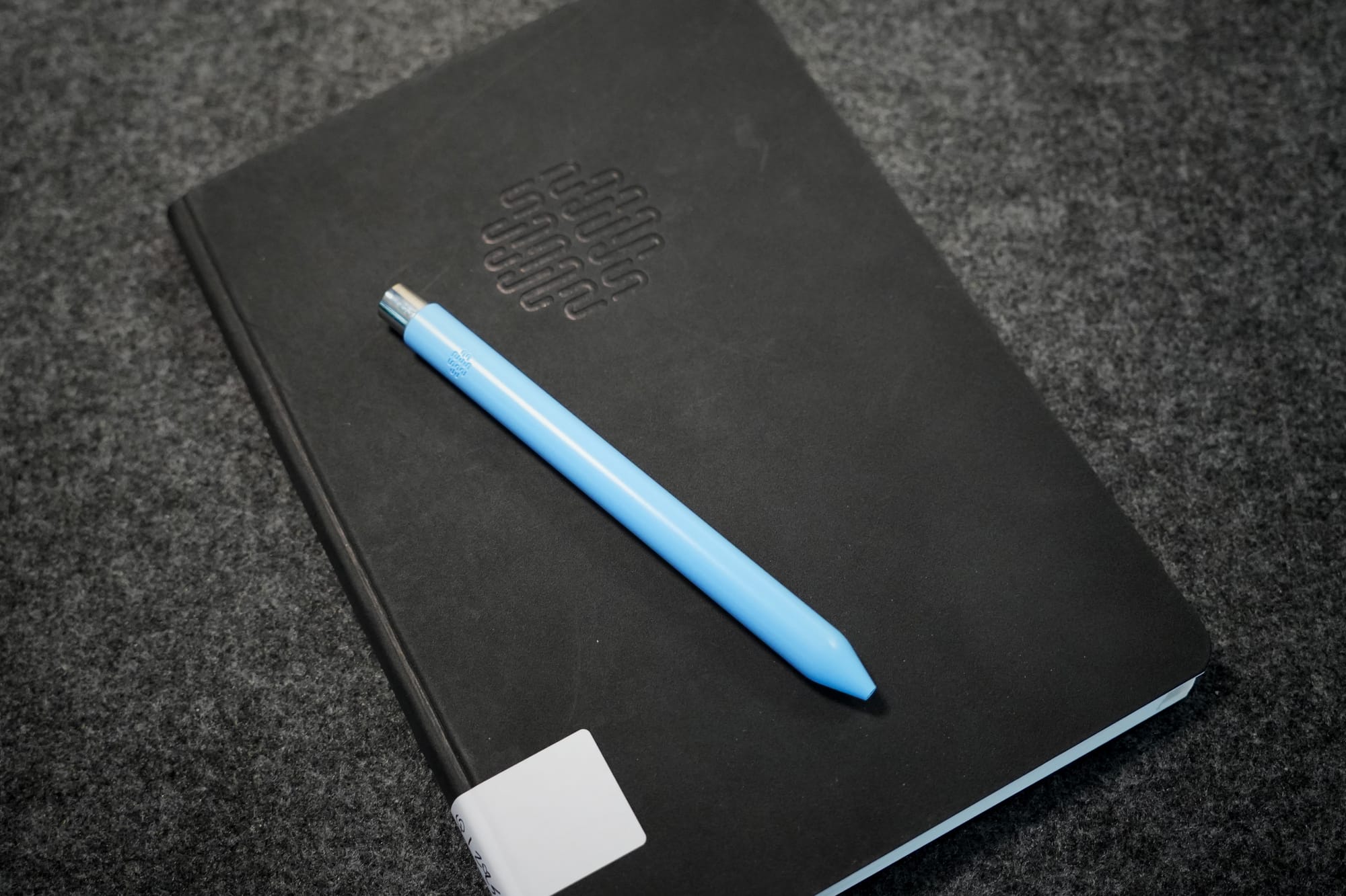 A closed black notebook with an embossed pattern on the cover, lying on a gray surface. A light blue pen rests diagonally across the notebook.