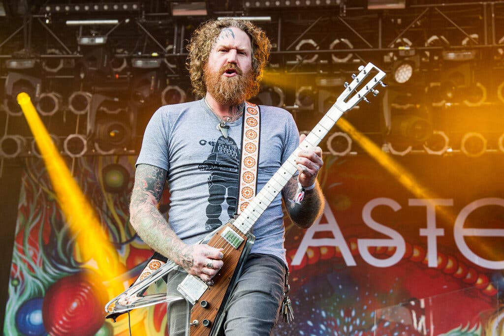 Brent Hinds of Mastodon performs live on stage, playing a Flying V guitar, with stage lights and the band’s colorful backdrop behind him.