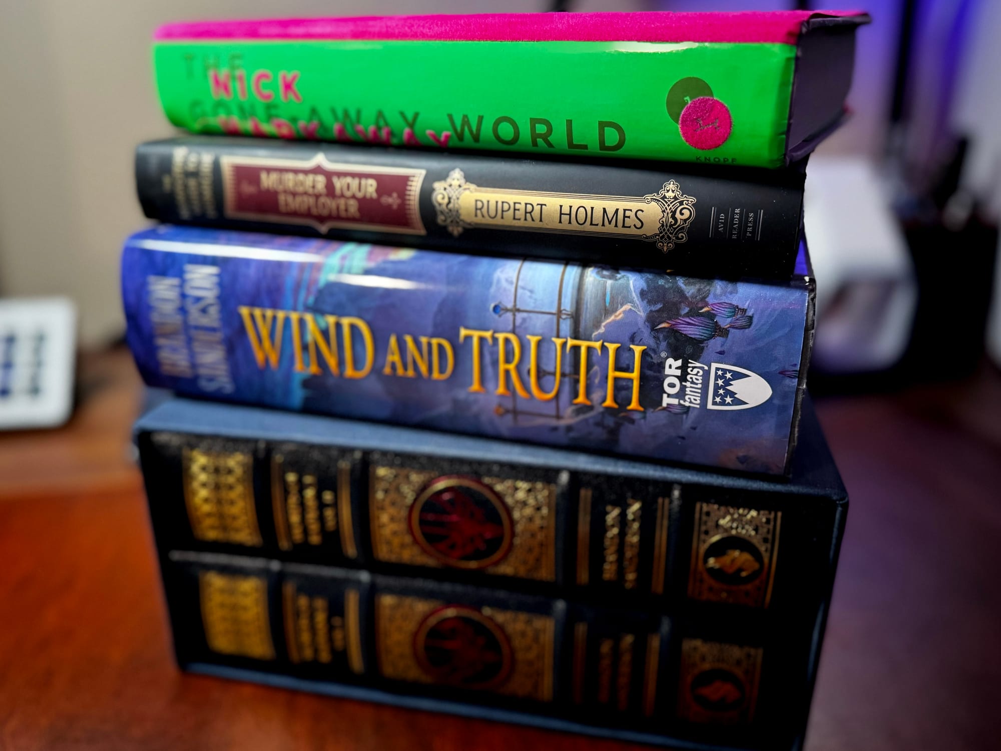 A small stack of books on a wooden surface. From top to bottom: The Gone-Away World by Nick Harkaway (bright green cover), Murder Your Employer by Rupert Holmes (black spine with ornate gold design), Wind and Truth by Brandon Sanderson (blue fantasy cover, Tor Fantasy), and two leatherbound Brandon Sanderson volumes stacked horizontally at the bottom.