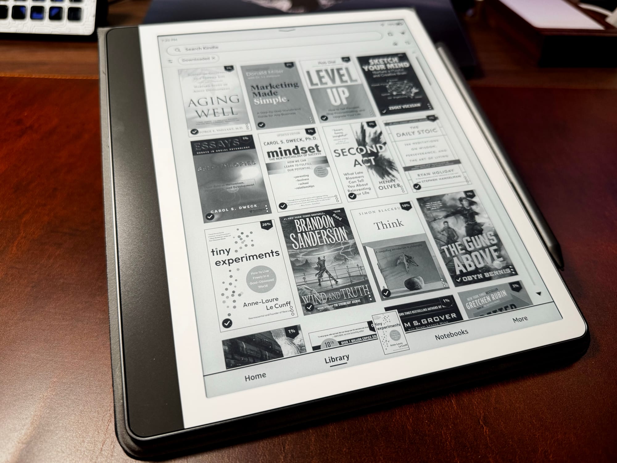 An e-reader on a desk displaying a Kindle library in black and white, with covers including Level Up, The Daily Stoic, Wind and Truth, and Tiny Experiments.