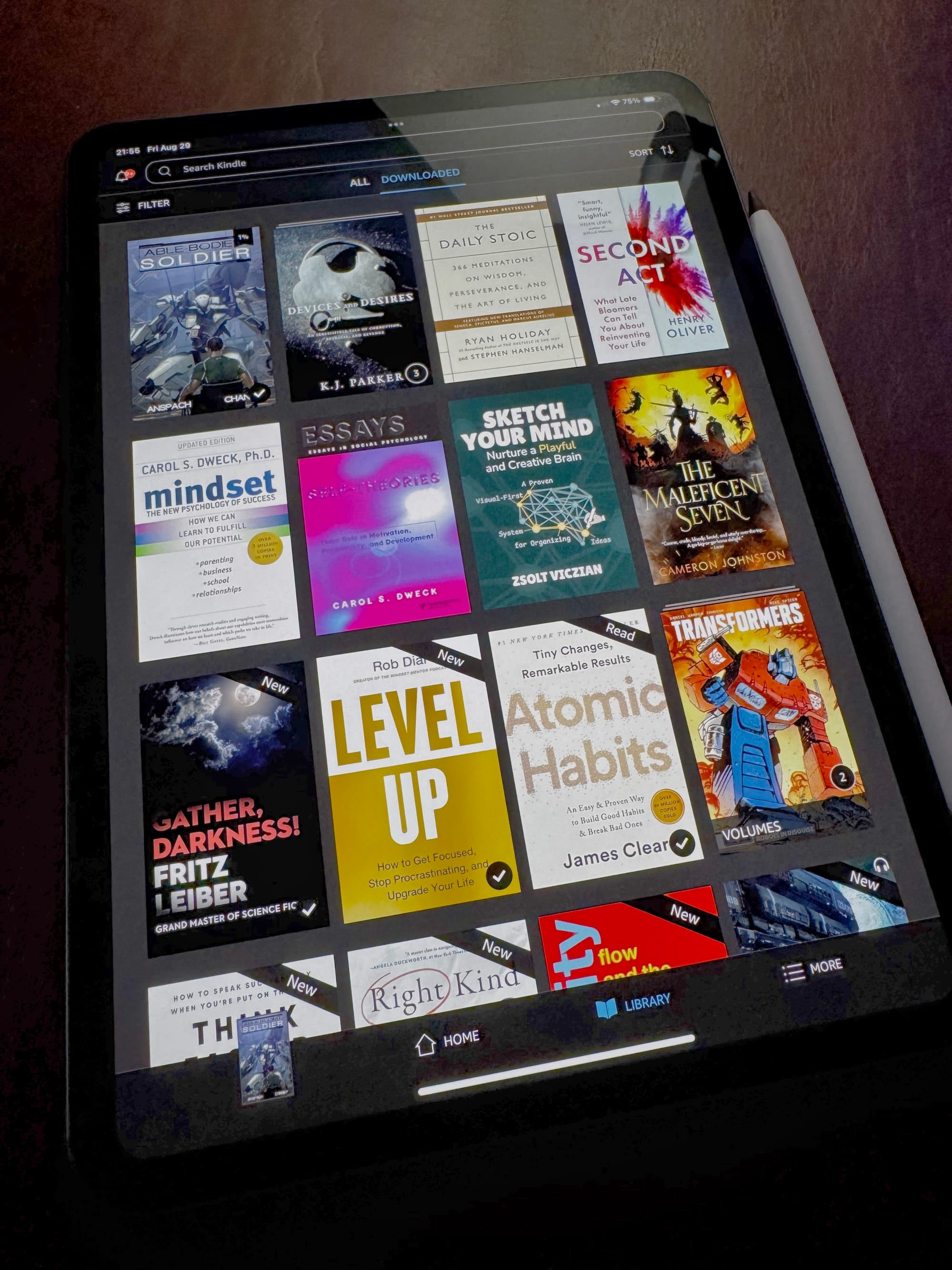 A tablet screen displaying a Kindle library with multiple book covers, including Atomic Habits, Mindset, The Daily Stoic, and others.