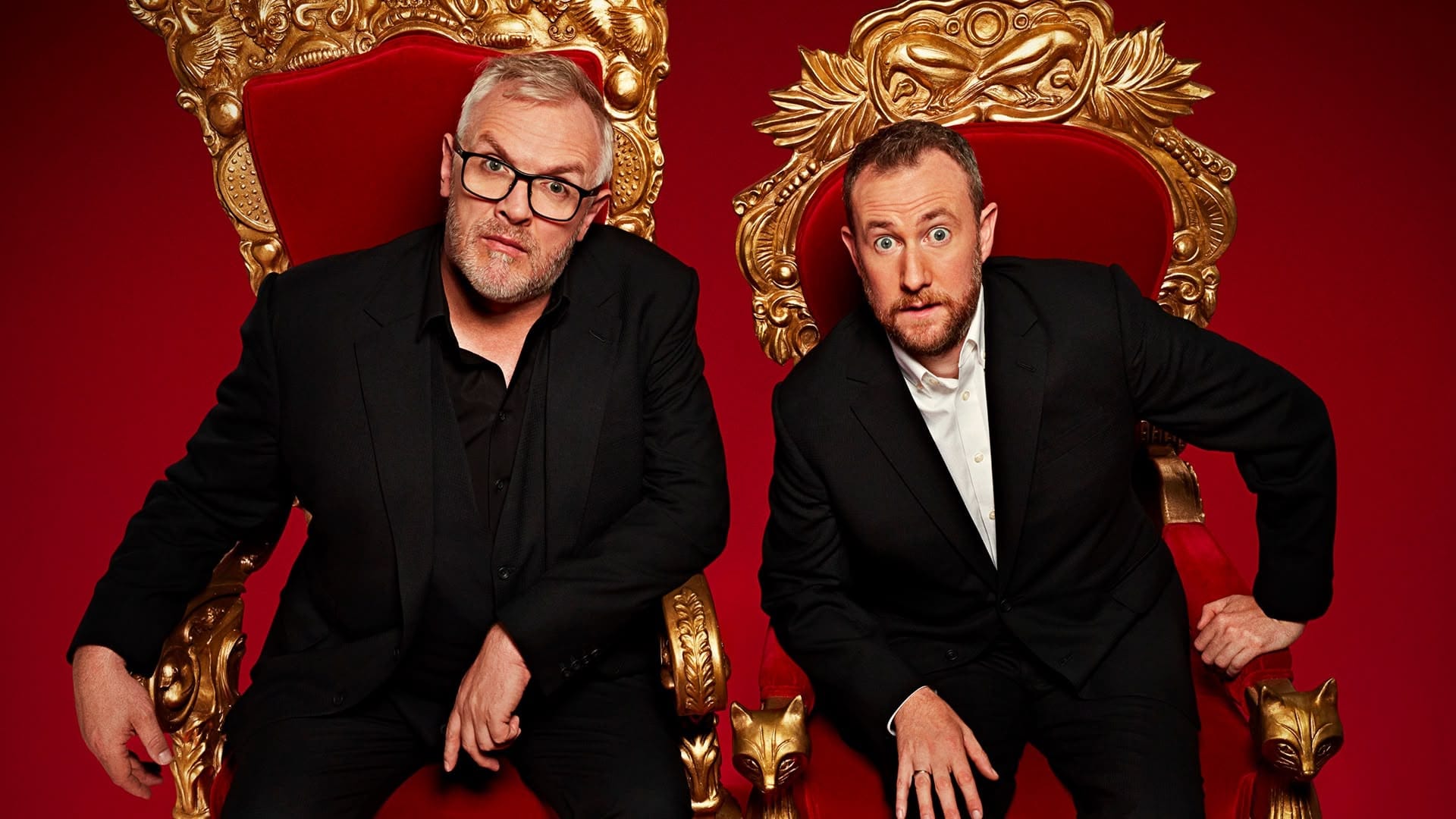 Comedians Greg Davies and Alex Horne, hosts of Taskmaster, sit in black suits on ornate golden thrones with red cushions, against a deep red background.