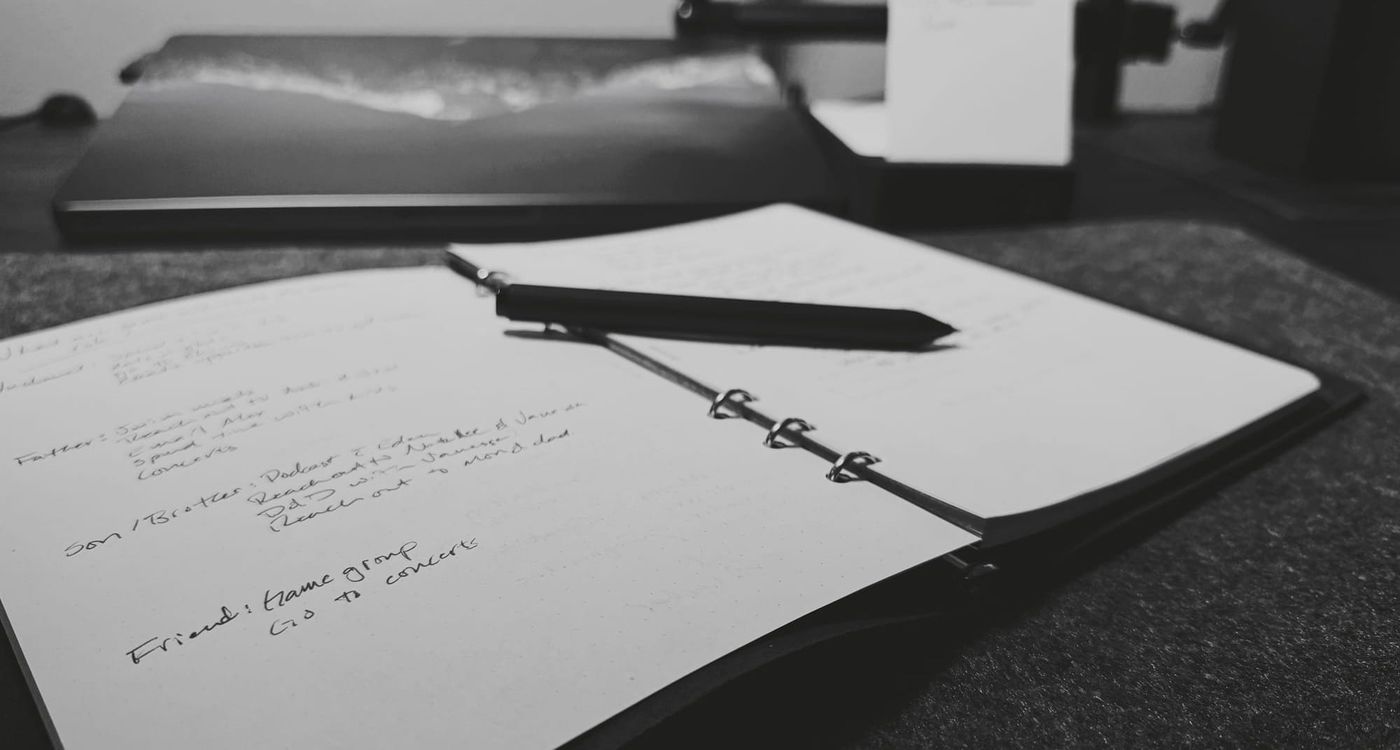 An open notebook with handwritten notes, lying on a desk. A black pen rests across the center of the notebook. In the background, a blurred laptop and a small notepad are visible.