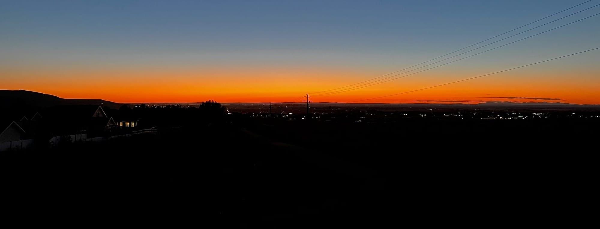 Twilight landscape with gradient sky from blue to orange, silhouetted houses and hills below, scattered lights visible.