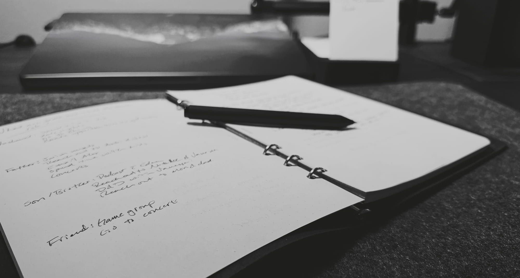 An open notebook with handwritten notes, lying on a desk. A black pen rests across the center of the notebook. In the background, a blurred laptop and a small notepad are visible.