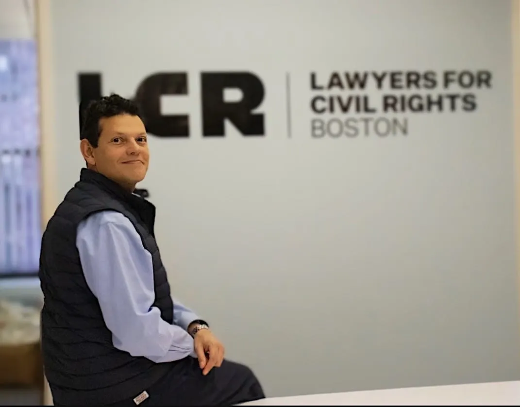 Iván Espinoza-Madrigal sentado frente al cartel de Abogados por los Derechos Civiles de Boston. Su oficina está ubicada en 61
