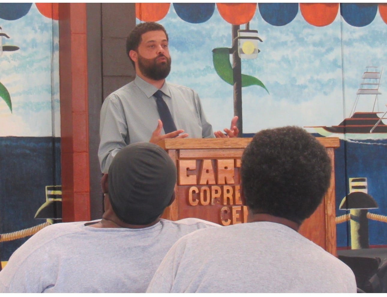 Jamir Jumoke speaks from a wooden podium at Carteret Correctional Center, gesturing with both hands. Two men are seated with their backs to the camera in the foreground. A painted harbor mural covers the wall behind him.