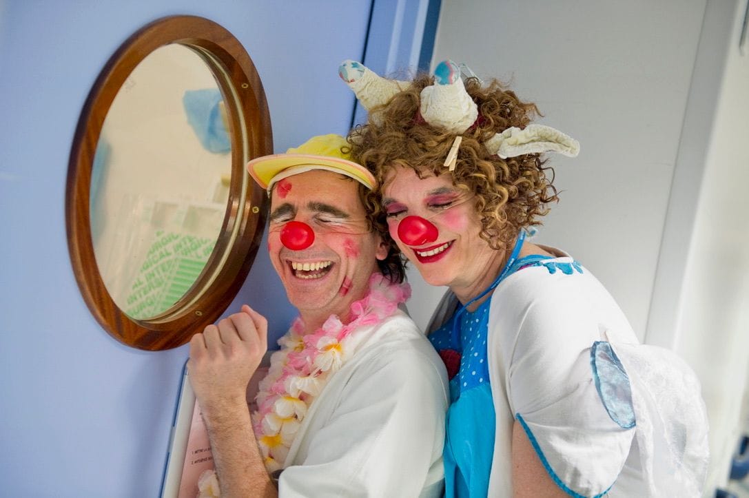 Two performers in white coats and red noses lean together against a hospital wall beside a round porthole window, eyes closed, laughing. One wears a yellow visor and a pink flower lei, the other a pale giraffe headpiece.