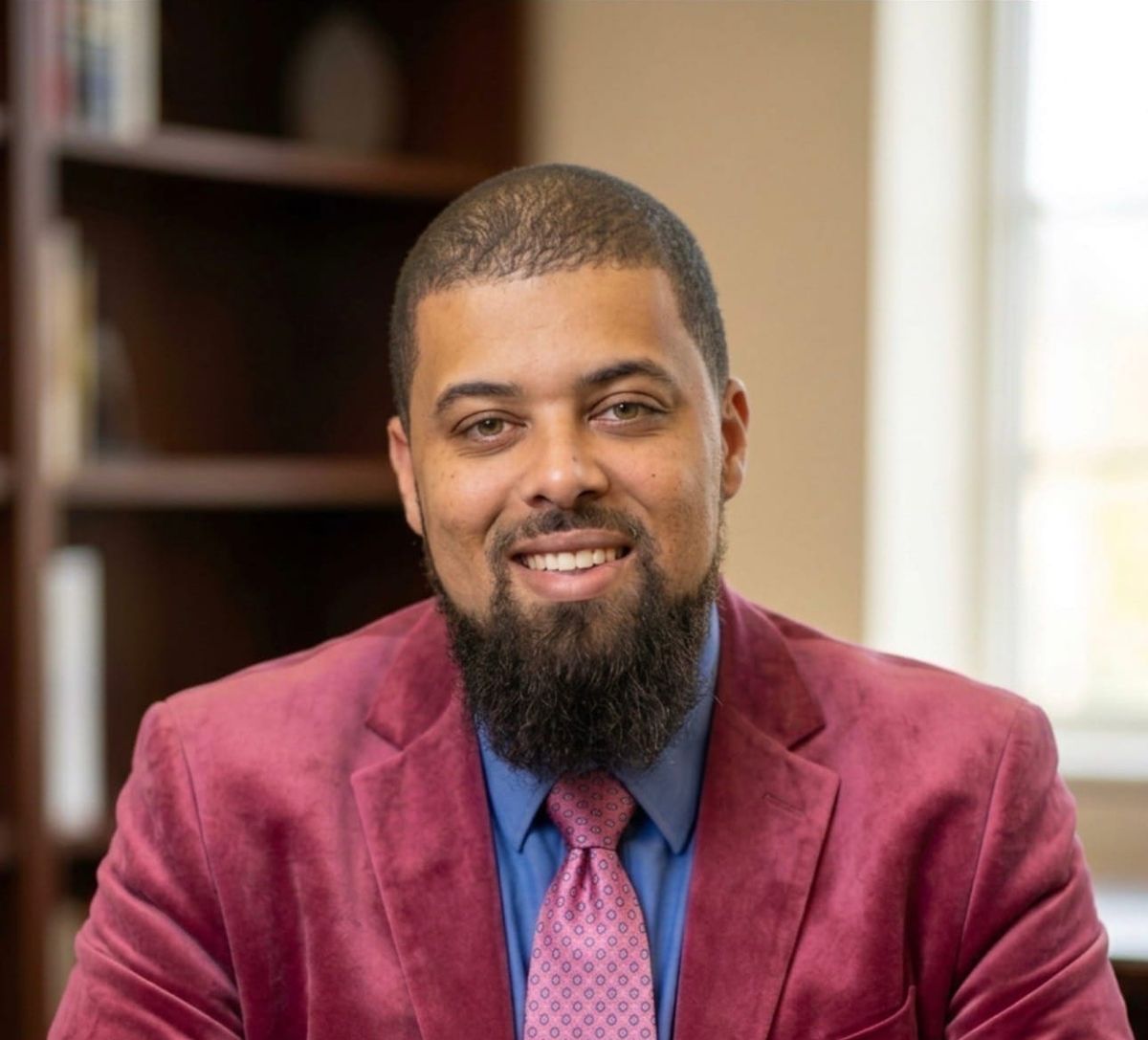 Jamir Jumoke, seated indoors with hands clasped, wearing a pink shirt over a blue collared shirt.