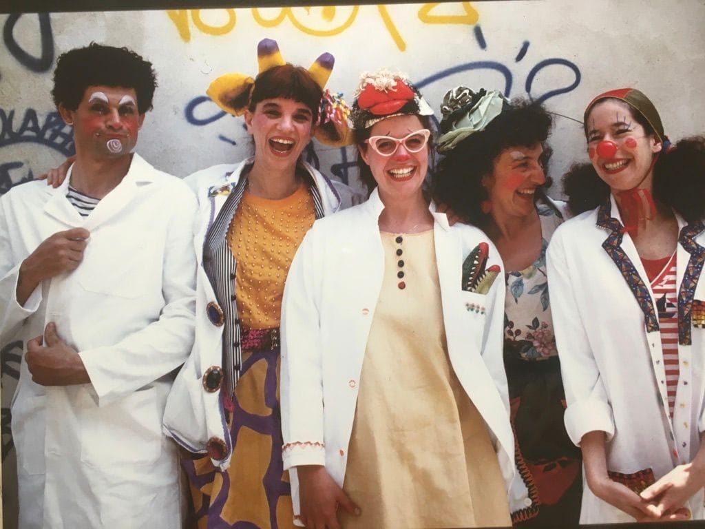 Five performers in white medical coats worn over brightly coloured costumes, laughing together in front of a graffitied wall. Several wear red noses, painted cheeks, and improvised headpieces
