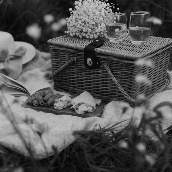 A picnic baskit sits with white flowers on a blanket near a plate of meat and cheese