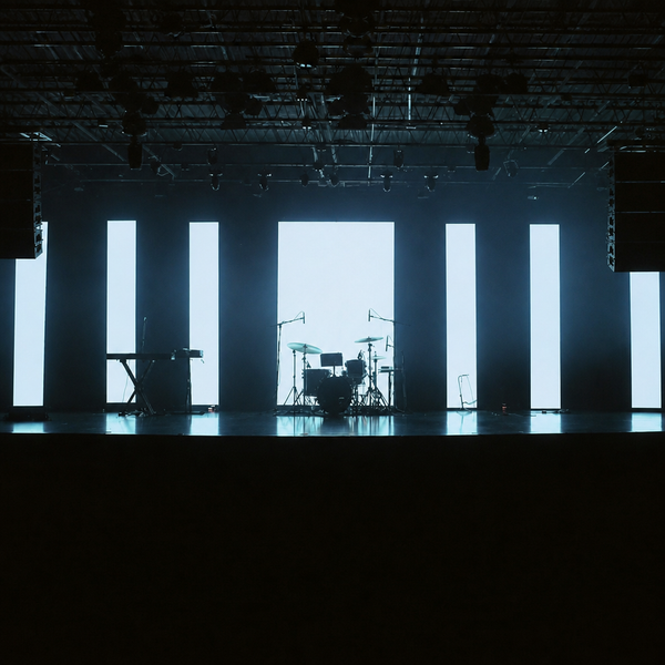 Empty concert stage backlit by tall white panels, a drum kit and keyboard standing unplayed under exposed lighting truss.