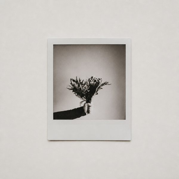A monochrome instant photograph on a pale wall, showing a hand holding out a small bouquet of flowers.